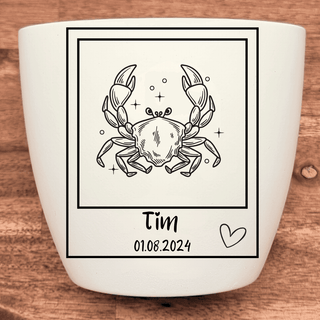 Nahaufnahme eines gravierten Blumentopfs mit Sternzeichen Krebs-Motiv, personalisiert mit Name 'Tim', Datum und Herzsymbol, auf Holzuntergrund.