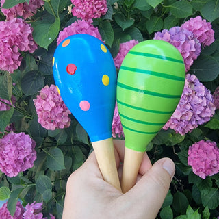 Hand hält zwei bunte Holz-Babyrasseln (Maracas) von Relaxday vor blühenden Hortensien. Ideal zum Greifen und Musizieren.