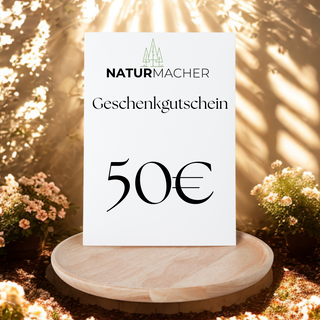 Naturmacher Geschenkgutschein im Wert von 50€ auf Holzteller, umgeben von Pflanzen im Sonnenlicht.