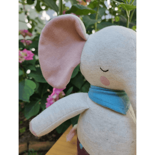 Ava&Yves Kuschelelefant aus Bio-Strick mit rosa Ohren und blauem Halstuch vor Pflanzen.