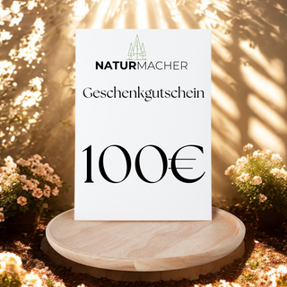 Naturmacher Geschenkgutschein im Wert von 100€ auf Holzteller, umgeben von Pflanzen.