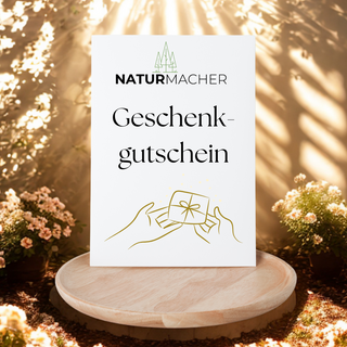 Naturmacher Geschenkgutschein: Weiße Karte mit Geschenksymbol und Naturmacher-Logo auf Holzteller im Garten.