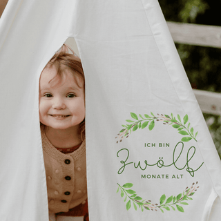 Baby schaut lächelnd hinter Meilensteinkarte "12 Monate" von Naturmacher hervor. Baby Meilensteinkarten fürs erste Jahr.