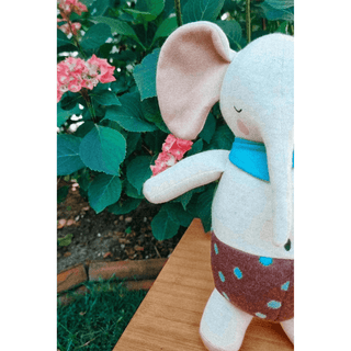 Kuscheliger Bio-Strick Elefant von ava&yves mit blauem Schal und gepunkteter Hose vor Pflanzen.