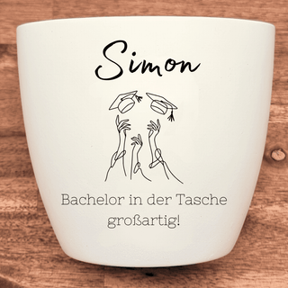 Weißer Blumentopf mit "Simon", geworfenen Doktorhüten und "Bachelor in der Tasche großartig!" graviert. Abschlussgeschenk.