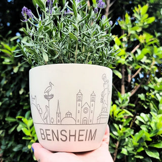 Keramiktopf mit Bensheim-Motiven und Lavendelpflanze, gehalten von einer Hand vor grünem Hintergrund.