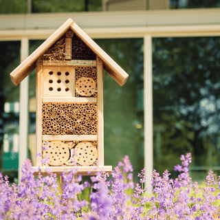 Insektenhotel aus Holz, gefüllt mit Naturmaterialien, vor violetten Blüten. Bauanleitung für Kinder von Naturmacher.