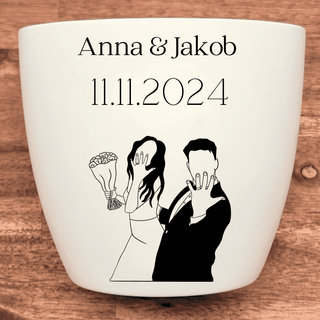 Weißer Blumentopf mit Gravur eines Brautpaares, das seine Ringe zeigt. Personalisiert mit "Anna & Jakob" und "11.11.2024"....