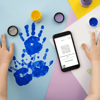 Babyhände mit blauer Farbe neben Smartphone mit Naturmacher Meilensteinkarten für Babys erstes Jahr. Download verfügbar.