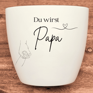 Weißer Blumentopf mit Gravur "Du wirst Papa" und Babyhand in Erwachsenenhand. Geschenk für werdende Väter.