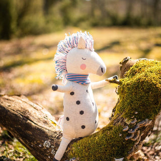 Ava&Yves Regenbogen-Pferd Kuscheltier aus Bio-Strick mit Punkten und buntem Schopf auf einem bemoosten Baumstamm.